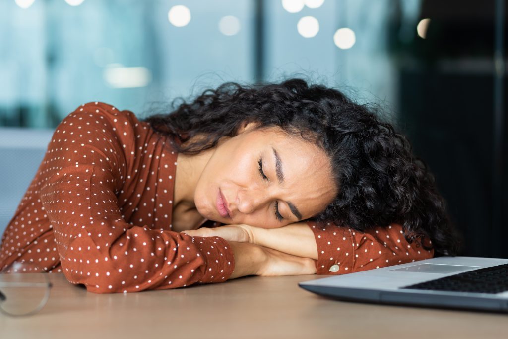 A woman with curly hair, wearing a brown polka dot shirt, rests her head on her crossed arms on a desk next to a laptop in this Sample Post.