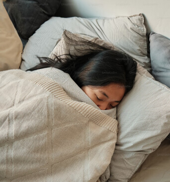 A person with long dark hair sleeps under a beige blanket, resting on striped pillows in a bed.
