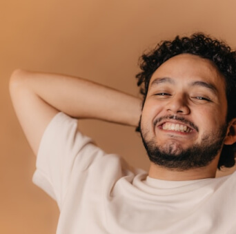A person with curly hair and a beard smiles while leaning back with hands behind their head, wearing a light-colored shirt against a warm, solid background.