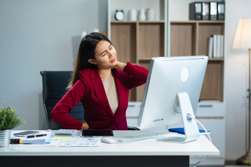 A woman in a red blazer sits at a desk in front of a computer, holding her neck and back in discomfort—perfect for illustrating a Sample Post about workplace ergonomics.
